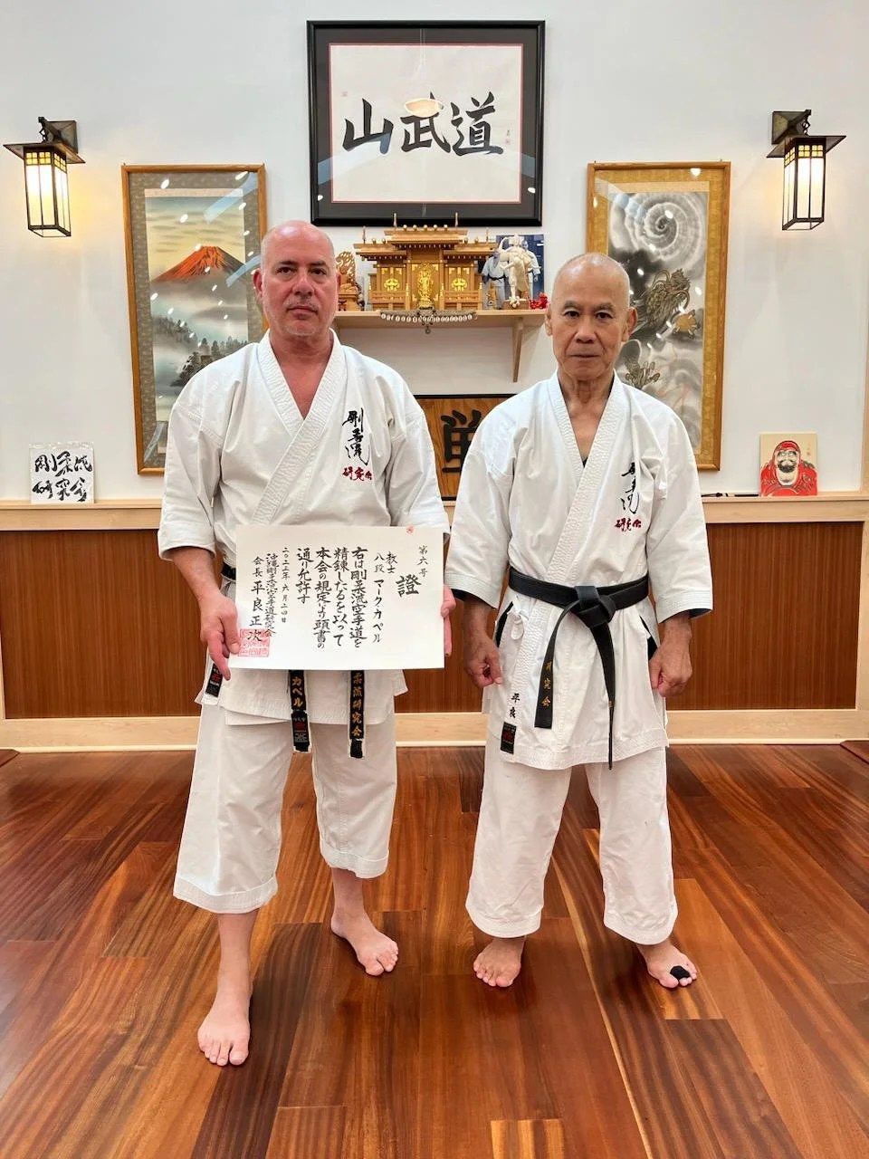 Sensei Kapel holds his Kyoshi 8th Dan certificate beside Masaji Taira Sensei in front of the shrine at the Holmdel dojo, under the framed 山武道 calligraphy.