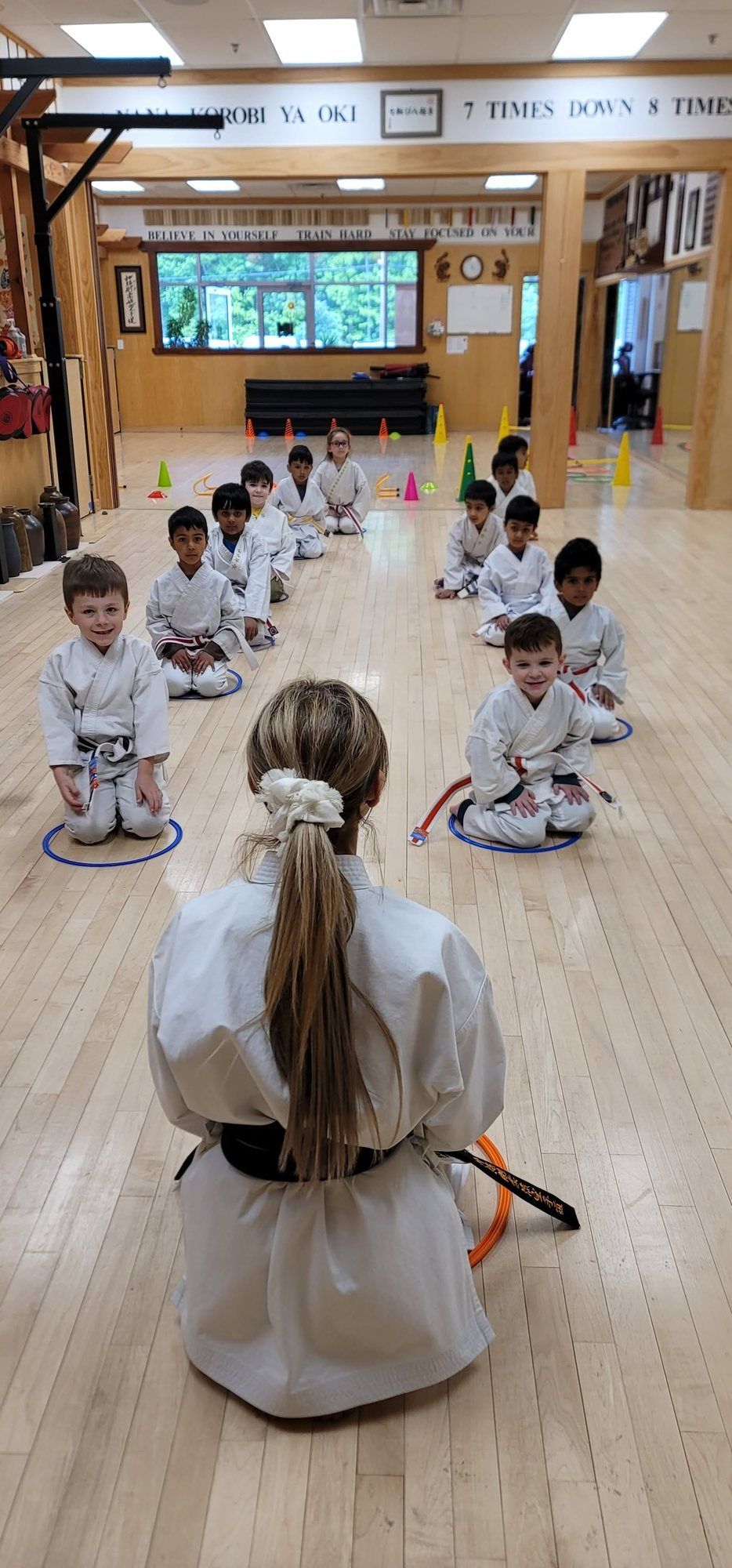 Young students in white gi kneel in a line on the Old Bridge dojo floor, facing their instructor.