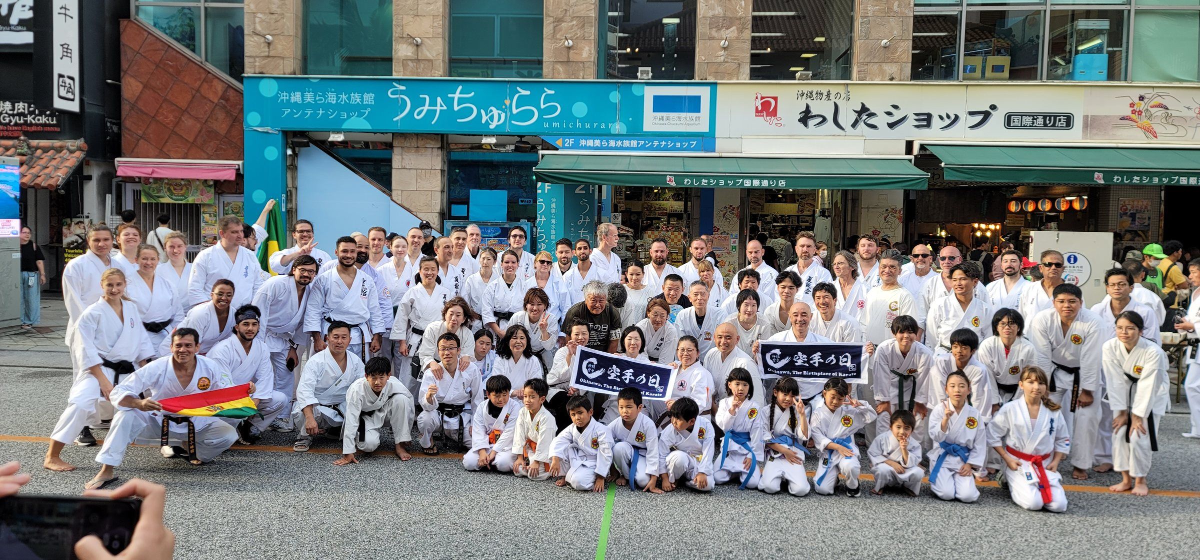 A large group of adults and children in white gi pose on a street in Naha, Okinawa, on Karate Day 2025.