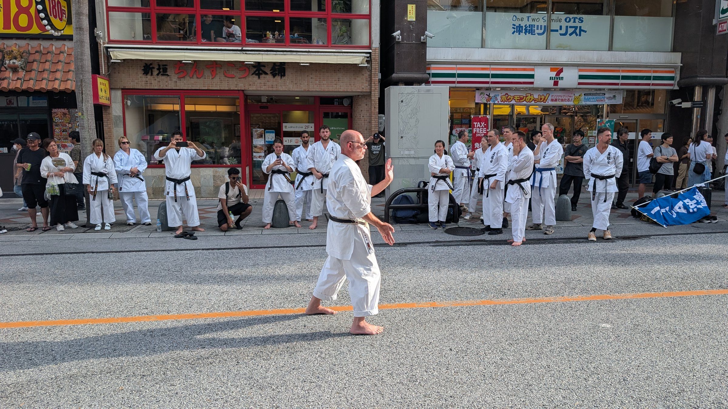 A black belt performs kata alone in the middle of the street on Kokusai-dori in Naha while others watch from the sides.