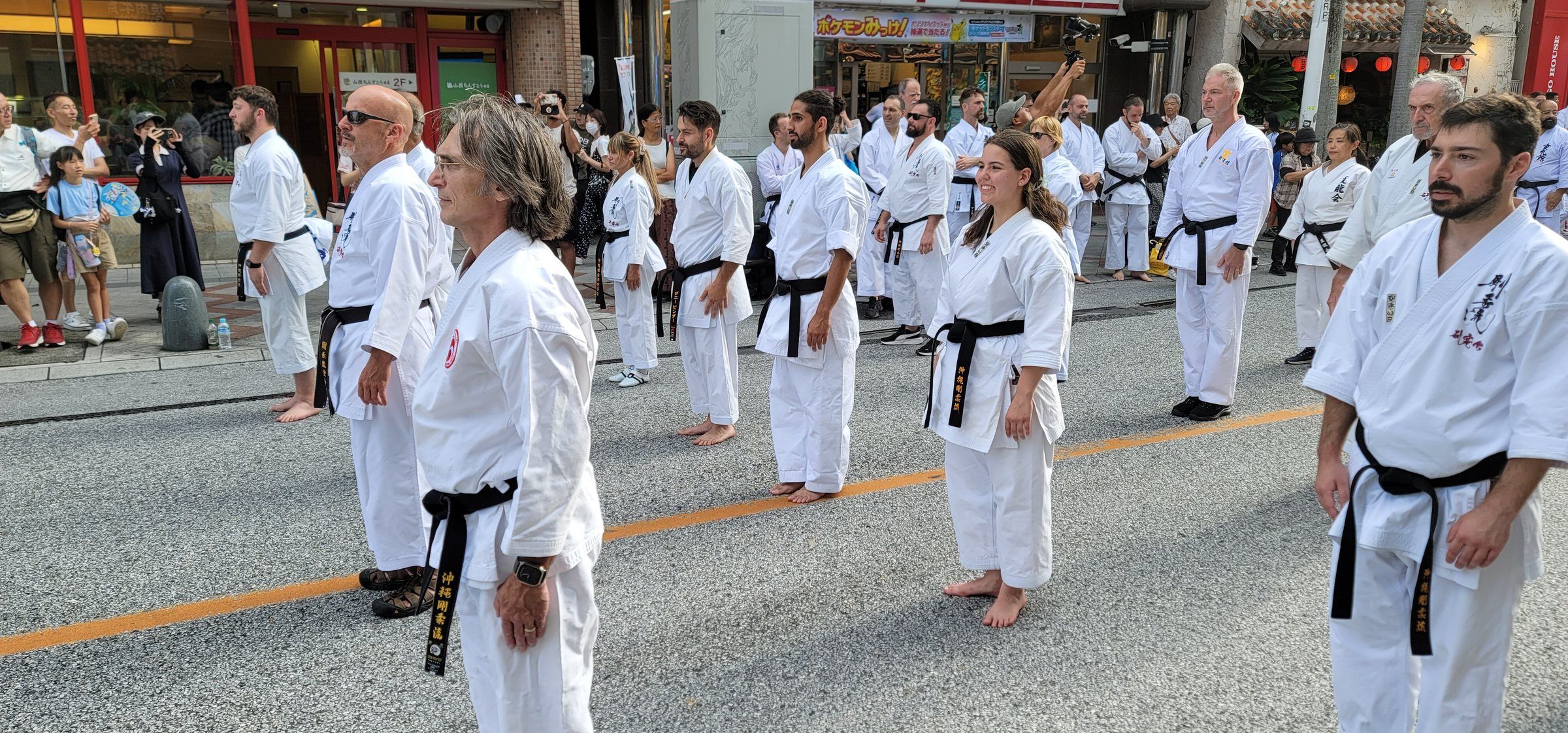 Karateka in white gi stand in lines on Kokusai-dori in Naha, waiting to perform kata on Karate Day 2025.