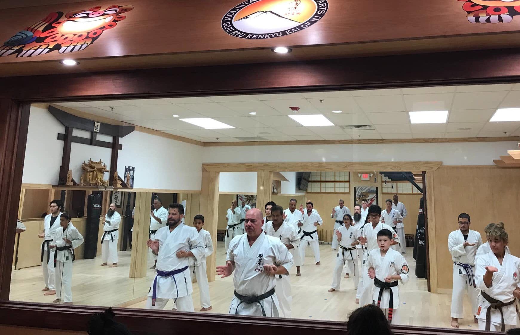 A class of adults and kids in white gi training at the Old Bridge dojo, seen through the front window.